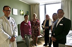 Eine Gruppe von Menschen, darunter ein Arzt und ein Pfarrer, stehen in einem hell eingerichteten Raum mit Bett und Sessel.